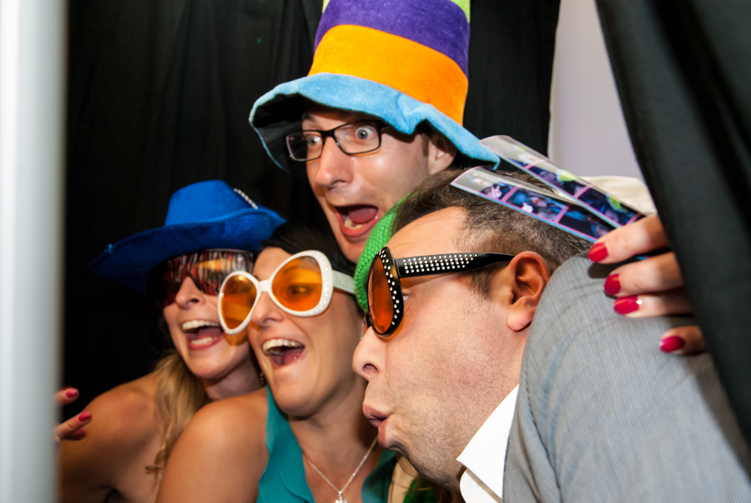 Wedding guests laughing in a closed photo booth wearing oversized sunglasses and a colorful top hat with a custom photo strip in hand at a Connecticut wedding