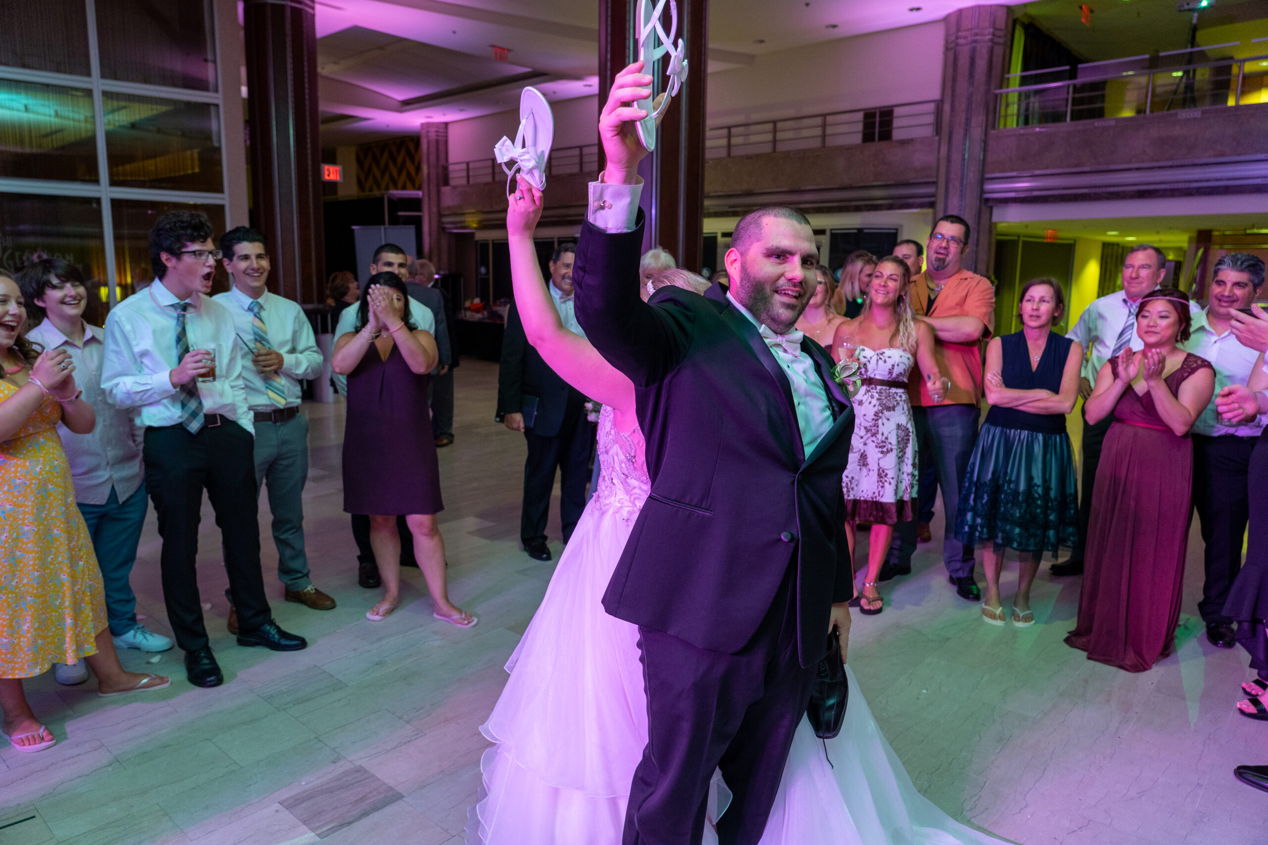 Groom raising his bride's shoes overhead as laughing guests circle the dance floor at a G. Fox Ballroom wedding reception in Hartford CT with purple uplighting