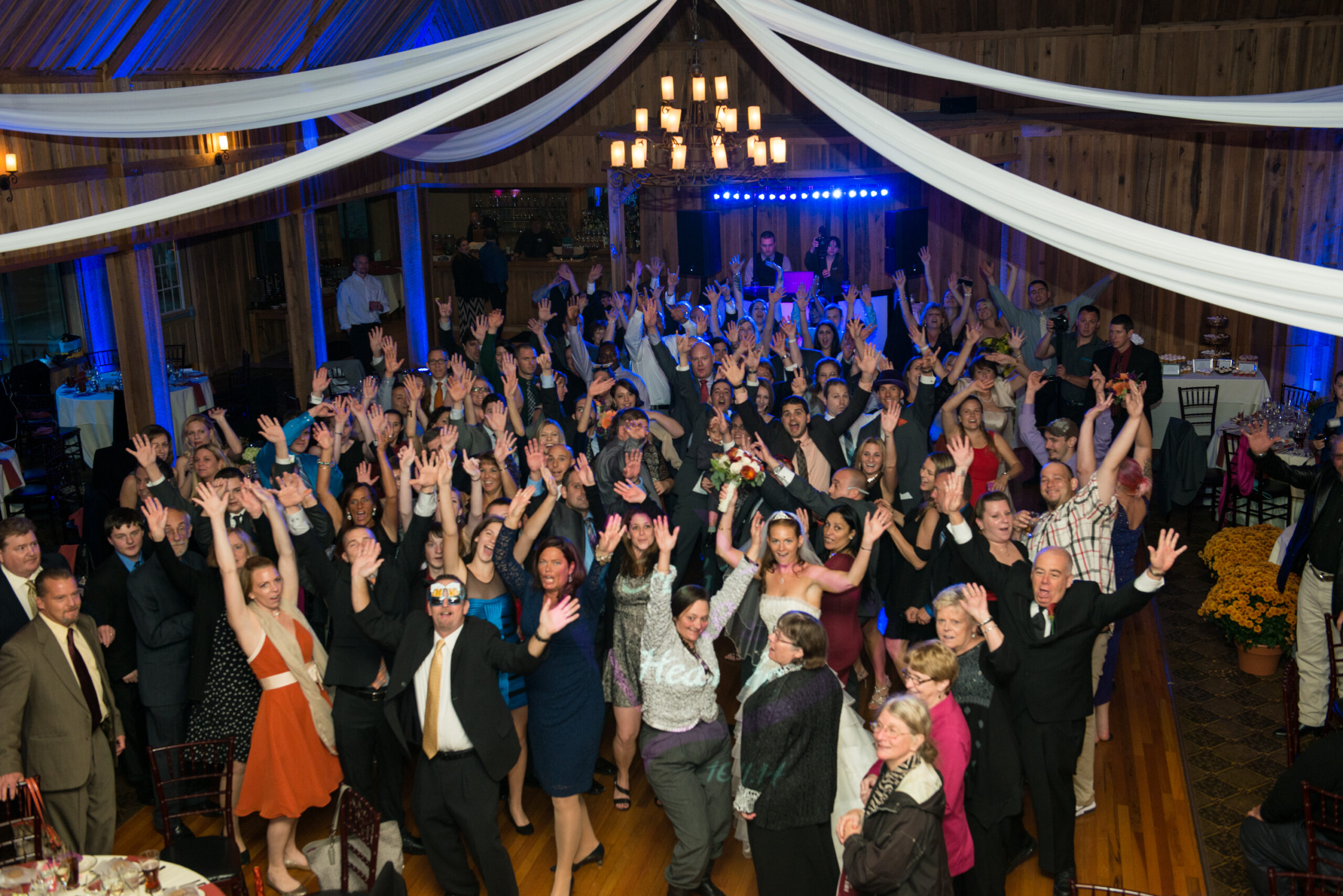 Entire wedding party and guests with hands raised on the dance floor at the Pavilion at Crystal Lake in Middletown CT with blue uplighting and rustic barn draping